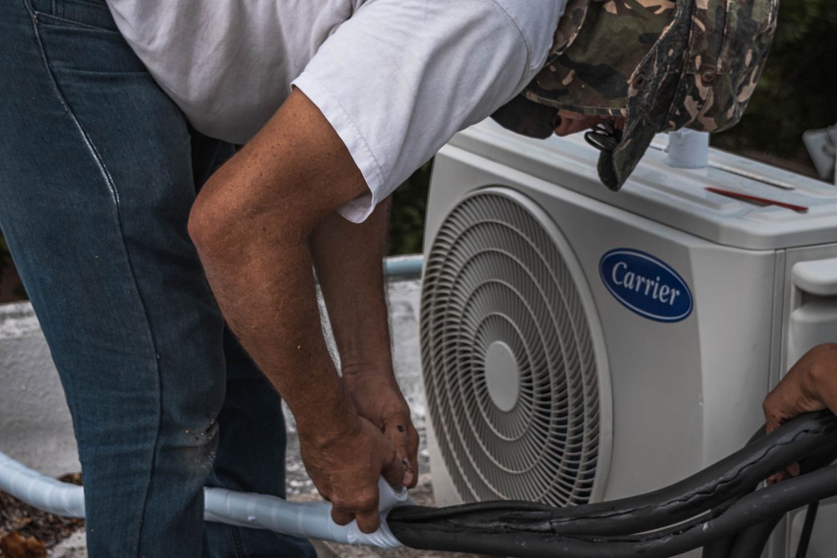 Technician checking hose and line connections on an outdoor AC unit during a maintenance visit.